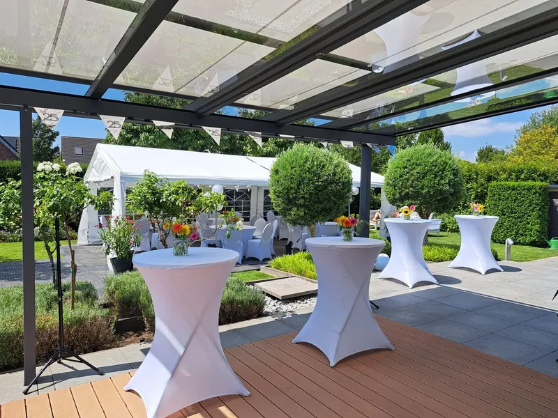 Stehtische mit weißen Hussen unter einer Terrassenüberdachung, dahinter ein weißes Partyzelt im Garten